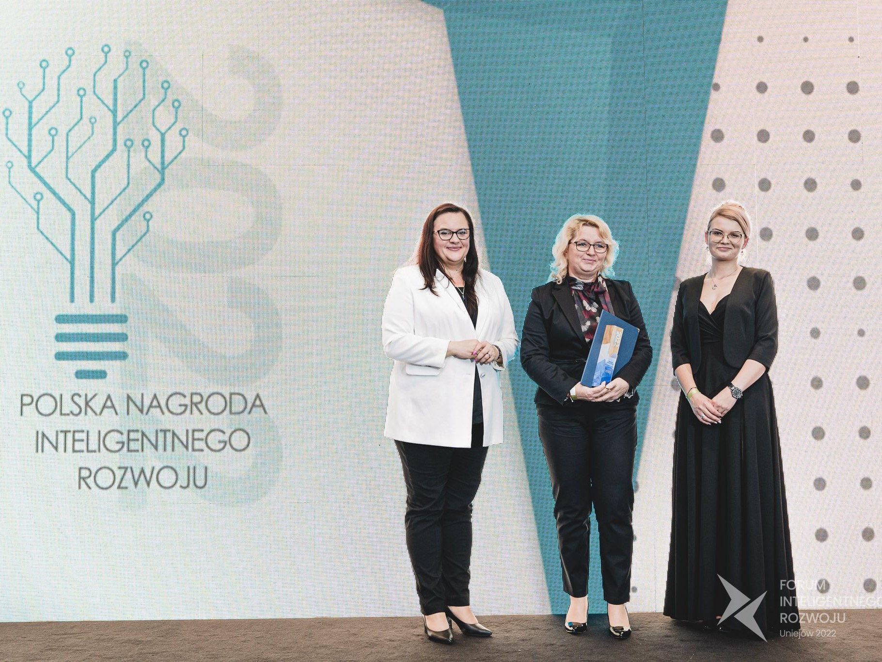Anna Mlost - Zastępca Dyrektora Departamentu Rozwoju Regionu wraz z dwiema kobietami stoi na scenie Forum Inteligentnego rozwoju po odebraniu nagrody