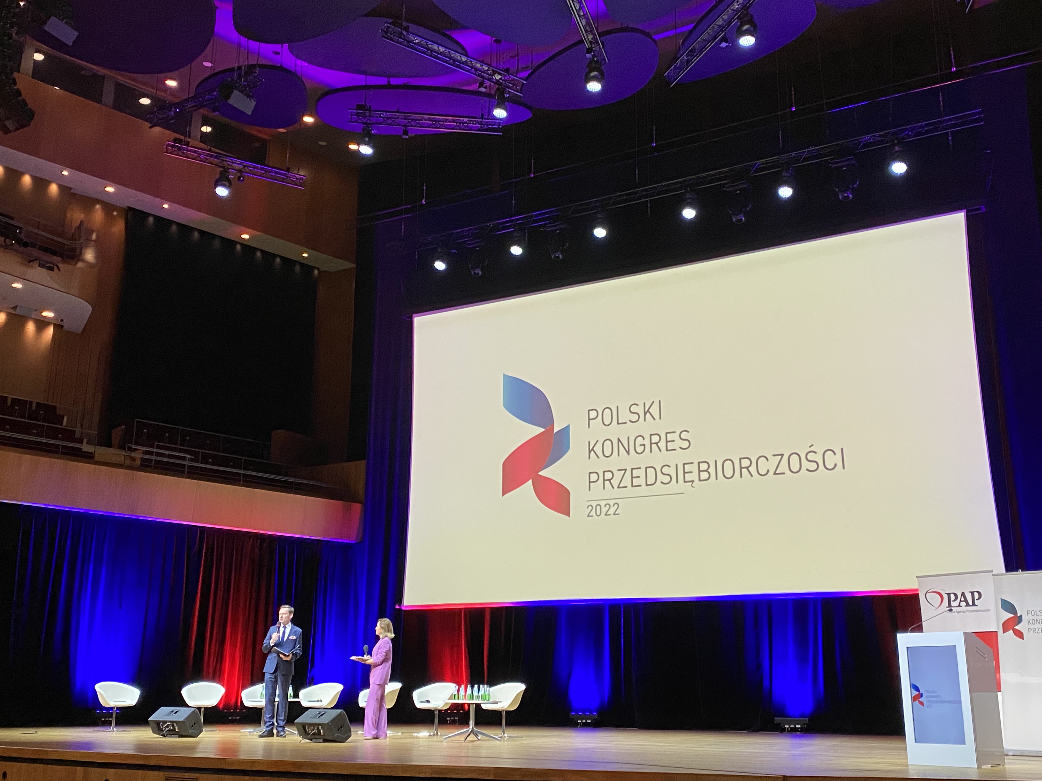scena oświetlona światłami niebieskim i czerwonym, w tle napis na ekranie &quot;Polski Kongres Przedsiębiorczości 2022&quot;. Na scenie dwoje prowadzących
