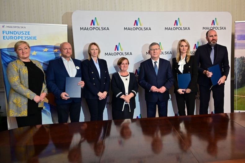 Zdjęcie przedstawia przedstawicieli Urzędu Marszałkowskiego Województwa Małopolskiego oraz przedstawicieli pośredników finansowych pożyczki REACT-EU na tle ścianki promocyjnej z logotypami Małopolski oraz rollupu z napisem Fundusze Europejskie