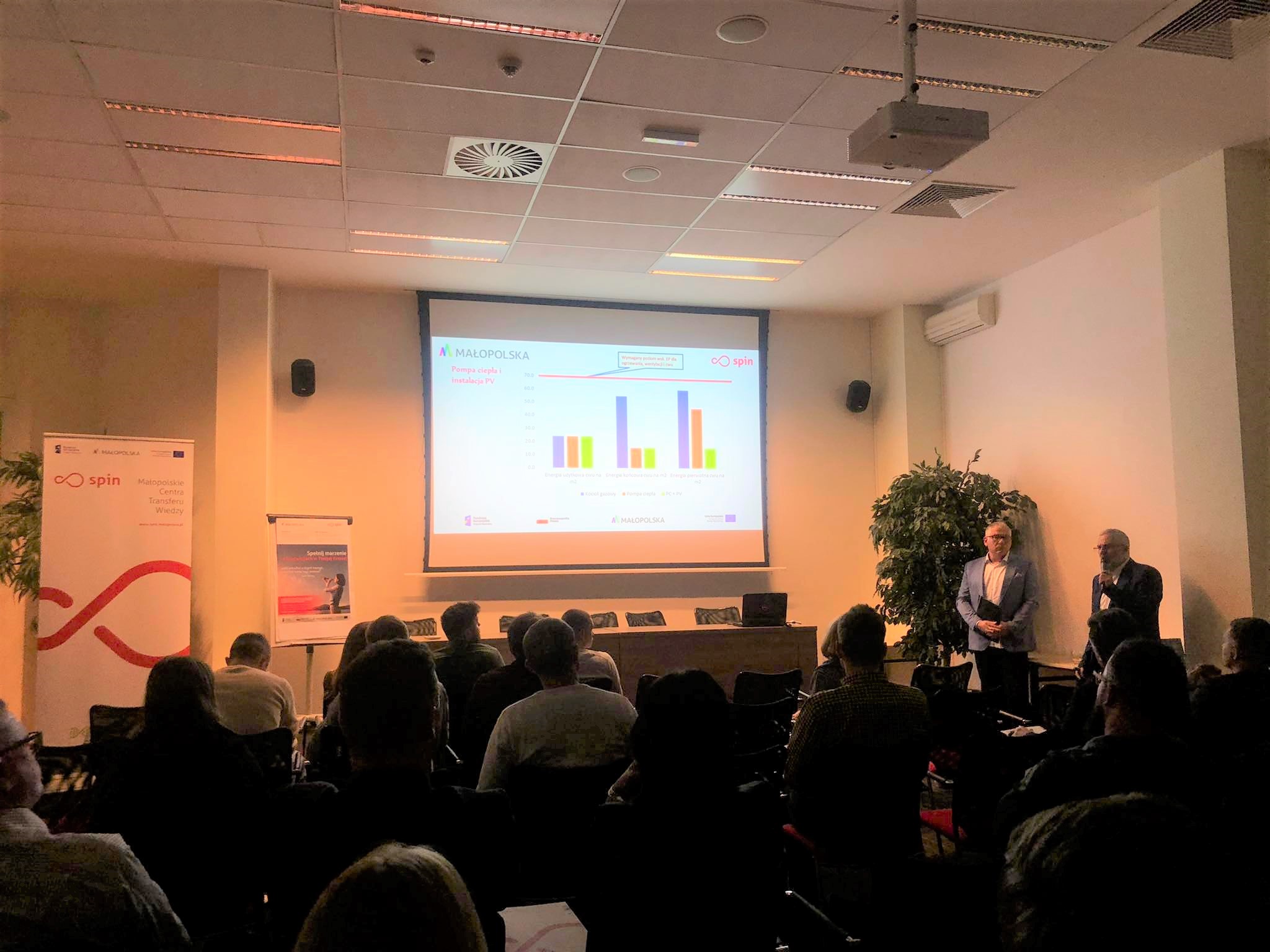 zdjęcie z konferencji SPIN CAMP. Publiczność,  na środku na ekranie dużego rzutnika wykresy, obok prelegent prowadzący konferencję