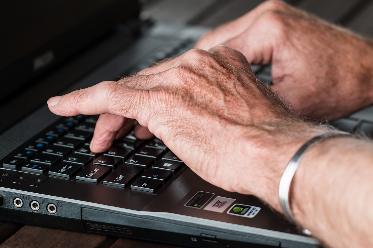 Grafika przedstawia ręce seniora piszącego na laptopie