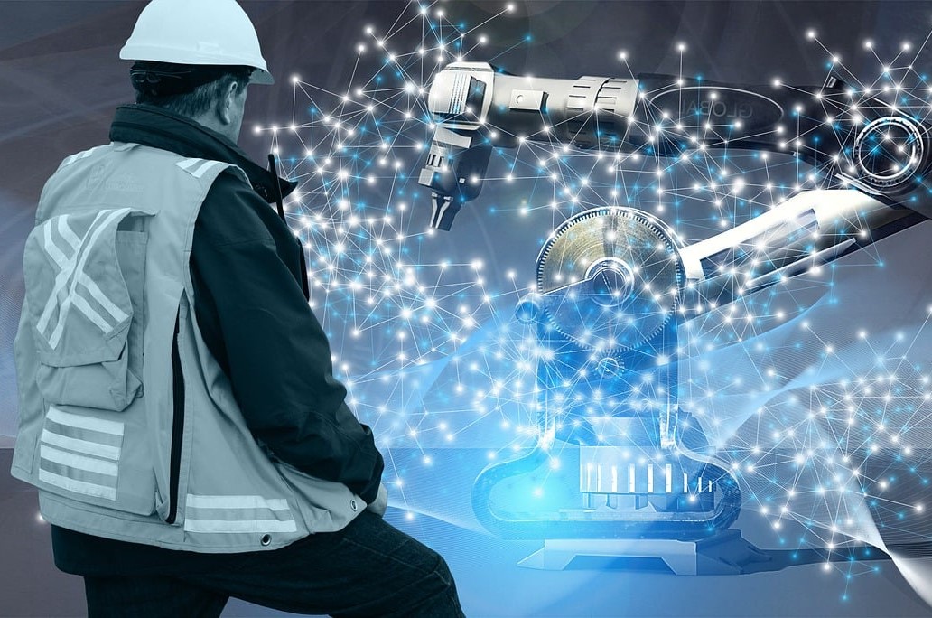Zdjęcie pracownika w kasku i kamizelce odblaskowej przyglądającego się z oddali pracy robota
