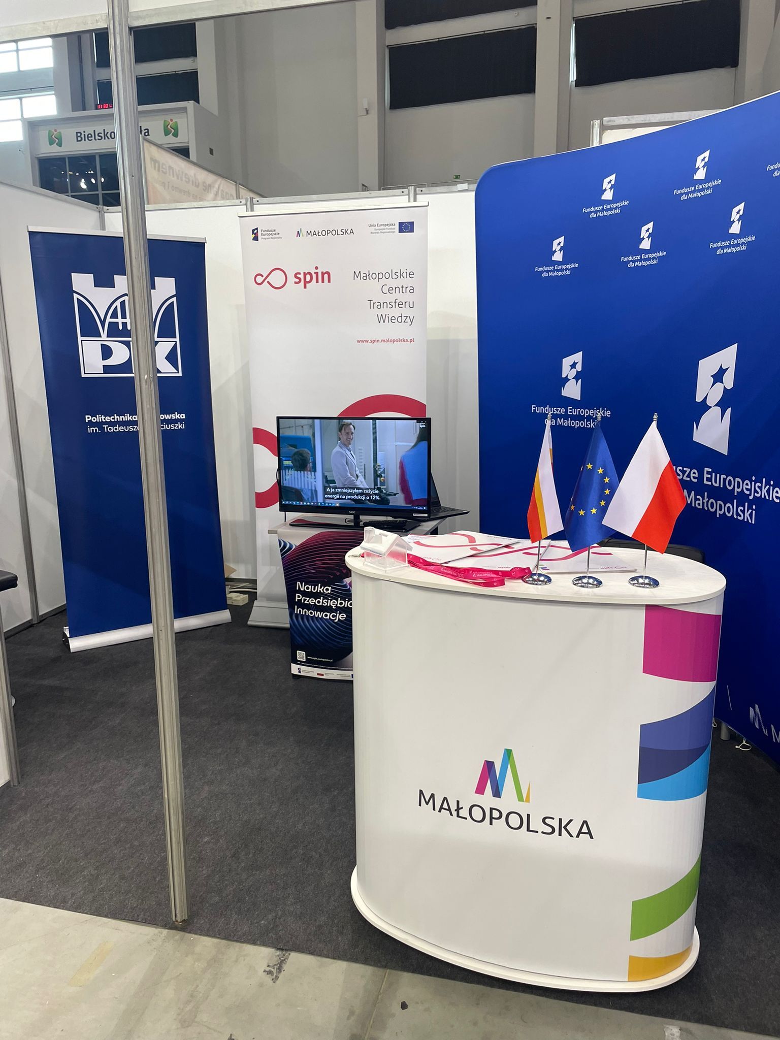 stoisko na targach w Bielsku Białej - stolik z flagami, telewizor oraz materiały ekspozycyjne typu rollup i ścianka